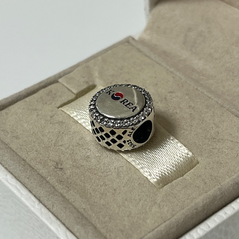May include: Silver charm with a round, black and white patterned design. The charm features a silver circle with a red, white, and blue Korean flag and the word "KOREA" in white lettering.