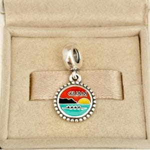 May include: A silver dangle charm featuring a circular pendant with a beaded edge. The centre displays an enamel scene of Guam with a red sky, a yellow sun, black mountains, and a turquoise ocean with a boat. The word GUAM is printed in white.