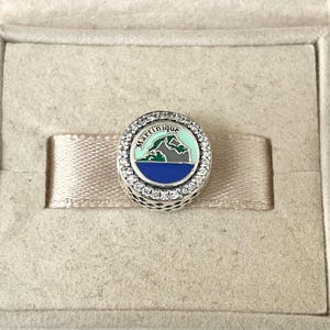 May include: Silver charm featuring a blue, green and grey enamel design of the island of Martinique. The charm is encircled by a ring of clear cubic zirconia stones. The word "Martinique" is written in white on the charm.