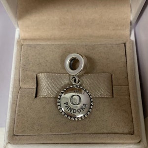 K&ouml;nnte beinhalten: Ein silberner Anh&auml;nger mit einem runden, strukturierten Design. Der Anh&auml;nger zeigt einen erhabenen Buchstaben "O" mit einer Krone dar&uuml;ber und dem Wort "PANDORA" darunter. Der Anh&auml;nger wird in einer wei&szlig;en Box mit beigefarbenem Innenraum pr&auml;sentiert.