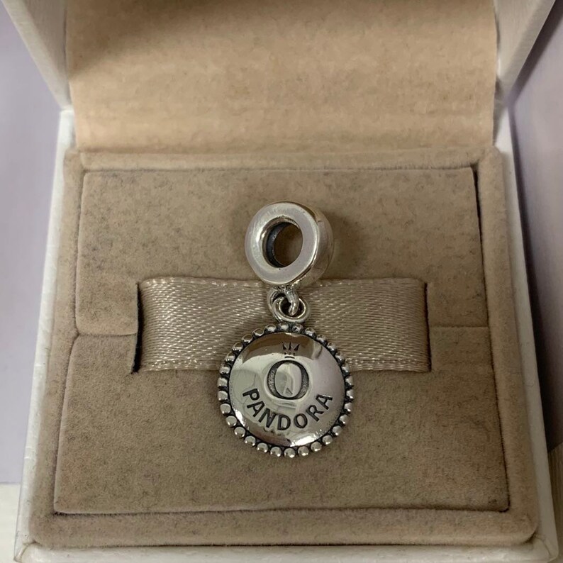 K&ouml;nnte beinhalten: Ein silberner Anh&auml;nger mit einem runden, strukturierten Design. Der Anh&auml;nger zeigt einen erhabenen Buchstaben "O" mit einer Krone dar&uuml;ber und dem Wort "PANDORA" darunter. Der Anh&auml;nger wird in einer wei&szlig;en Box mit beigefarbenem Innenraum pr&auml;sentiert.