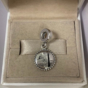 Puede incluir: Un charm de plata con una imagen en blanco y negro del Capitolio de los Estados Unidos y una bandera estadounidense. El charm est&aacute; en una caja blanca con un interior beige.