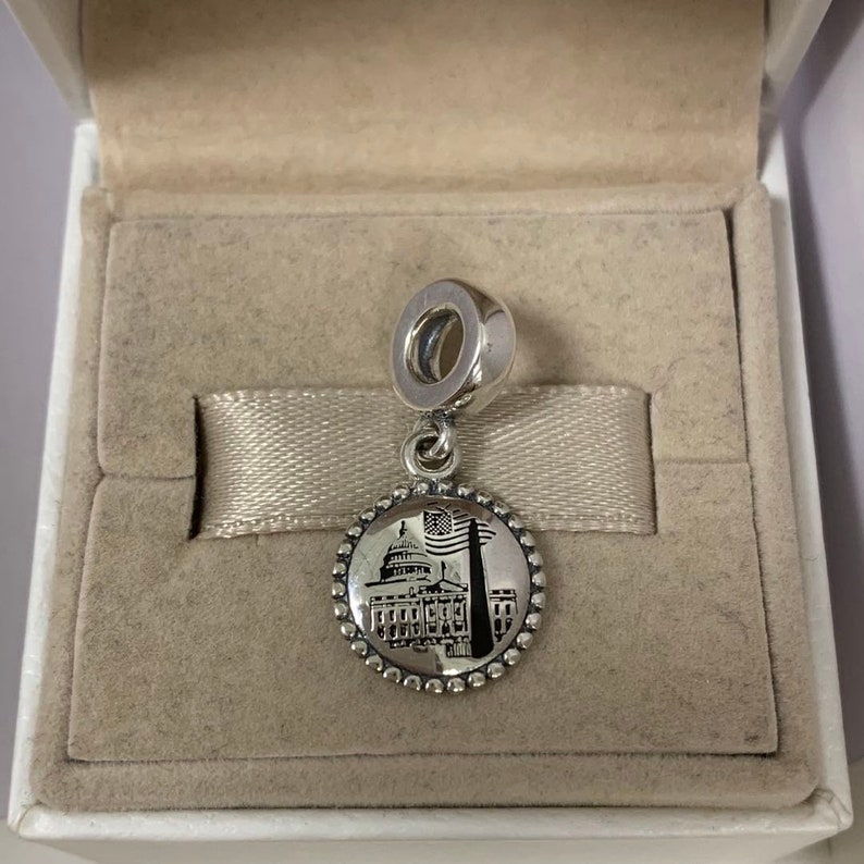 Puede incluir: Un charm de plata con una imagen en blanco y negro del Capitolio de los Estados Unidos y una bandera estadounidense. El charm est&aacute; en una caja blanca con un interior beige.