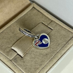 May include: Silver charm in the shape of a heart with a blue and red enamel design. The charm features a Union Jack flag and a crown with a blue enamel background. The charm is in a white box.