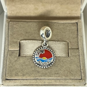 May include: A silver charm with a round pendant depicting a red and yellow sunset over a blue ocean with a palm tree. The word "Aruba" is written in blue. The charm is presented in a beige box.