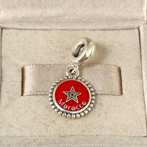 May include: A silver charm with a red enamel disc featuring a green star and the word "Morocco". The charm is round with a beaded edge and a loop for attaching to a bracelet or necklace. The charm is presented in a beige box.