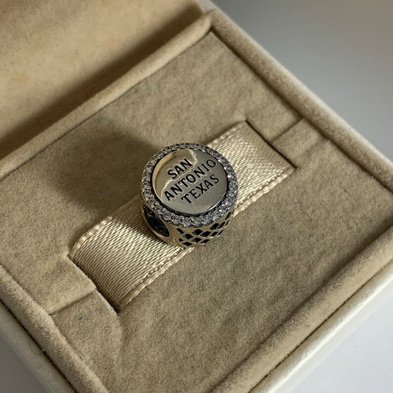 Puede incluir: Encanto de plata con un borde redondo de cristal transparente. El encanto presenta un disco de plata con el texto "SAN ANTONIO TEXAS" en letras negras.