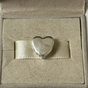 May include: A silver heart charm with an inscription around the edge. The charm is in a white box with a white ribbon.