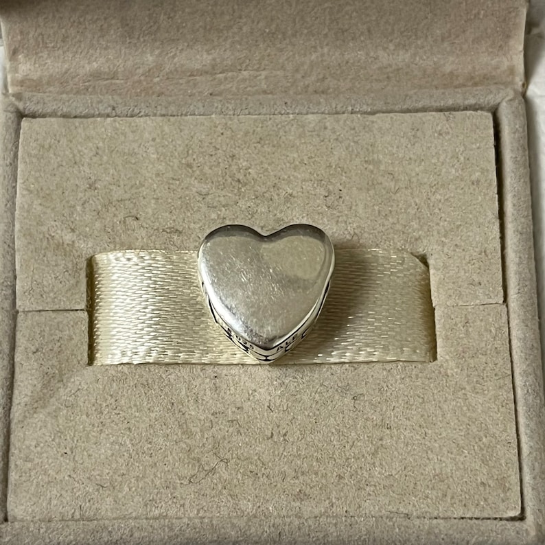 May include: A silver heart charm with an inscription around the edge. The charm is in a white box with a white ribbon.