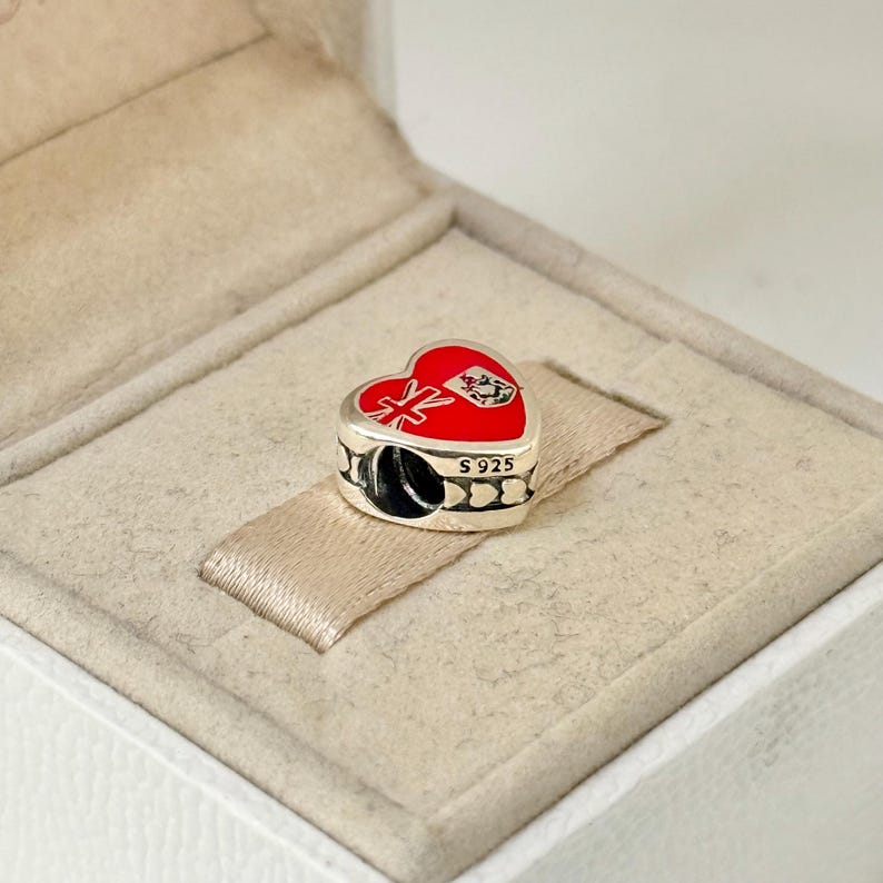 May include: A silver charm in the shape of a heart with a red enamel design featuring a cross and a crown. The charm has a small loop on the top and the words "S 925" on the side.