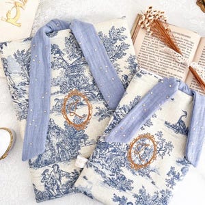 Peut inclure: Deux sacs fourre-tout à motif de toile de Jouy avec des anses bleu clair ornées de petits ornements scintillants. Les sacs présentent une scène pastorale bleue et blanche et une initiale brodée en or. Les sacs sont sur une surface en dentelle blanche.