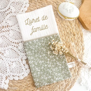 Peut inclure: Une couverture de livre en tissu blanc avec les mots "Livret de famille" brodés en or. La couverture a un motif floral vert sur la moitié inférieure.