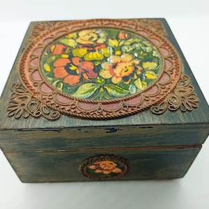 Puede incluir: Una caja de madera con un dise&ntilde;o floral pintado en la parte superior. La caja est&aacute; decorada con un borde de color cobre y tiene un acabado desgastado.