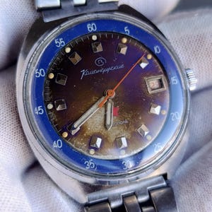 Puede incluir: Reloj de pulsera vintage con correa y caja de acero inoxidable. La esfera del reloj tiene un bisel azul, una esfera marrón y púrpura, y agujas plateadas. La esfera presenta el texto "Командирские" y "тополь". El reloj también tiene una pantalla de fecha.