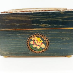 Puede incluir: Una caja de madera azul oscuro con un borde dorado y un dise&ntilde;o floral pintado en la parte delantera. La caja tiene una tapa con bisagras y un cierre de lat&oacute;n.