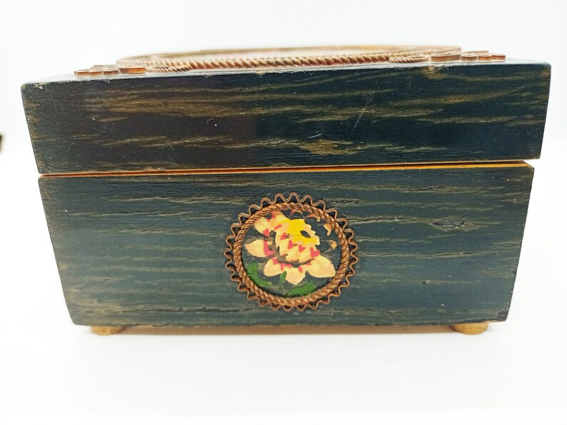 Puede incluir: Una caja de madera azul oscuro con un borde dorado y un dise&ntilde;o floral pintado en la parte delantera. La caja tiene una tapa con bisagras y un cierre de lat&oacute;n.