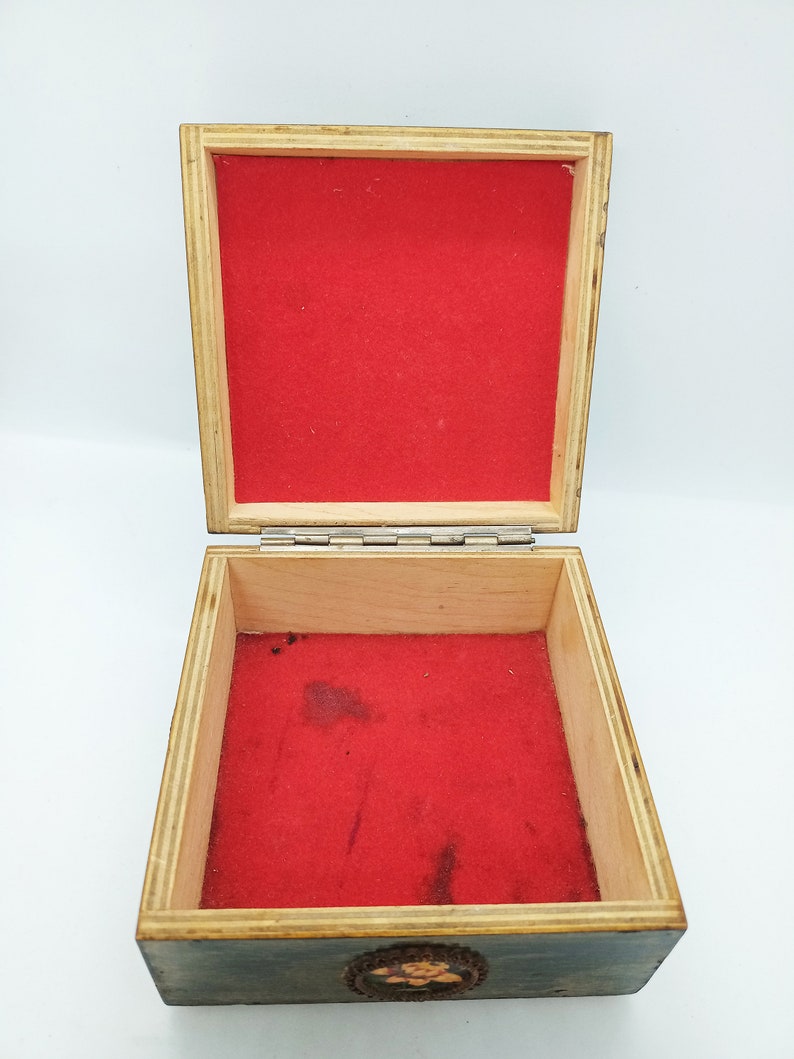 Puede incluir: Una caja de madera con tapa con bisagras y un interior de terciopelo rojo. La caja tiene un dise&ntilde;o decorativo en la parte delantera.