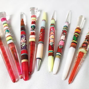 May include: A selection of decorative pens with clear, colourful, and patterned designs. The pens feature various colour schemes, including red, white, yellow, and floral accents. The pen tips are clear and the bodies are adorned with stripes and floral patterns.