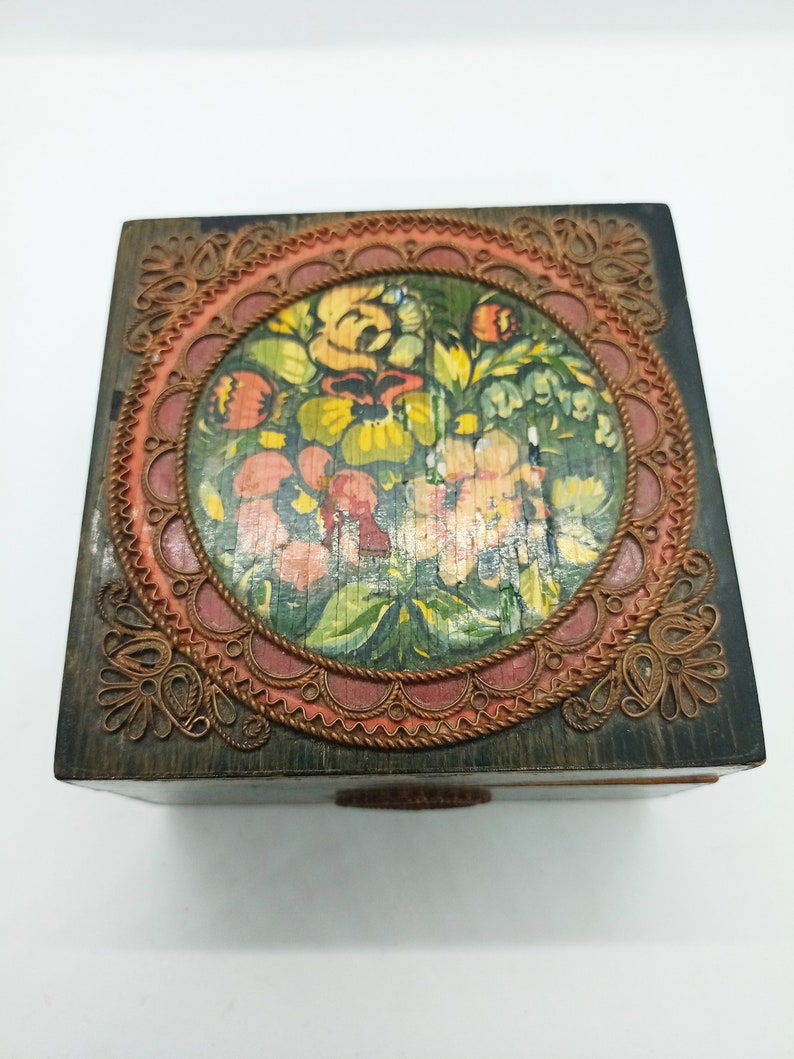 Puede incluir: Una caja de madera con un dise&ntilde;o floral pintado dentro de un marco circular. El marco est&aacute; decorado con un ribete tejido de cobre y rojo.