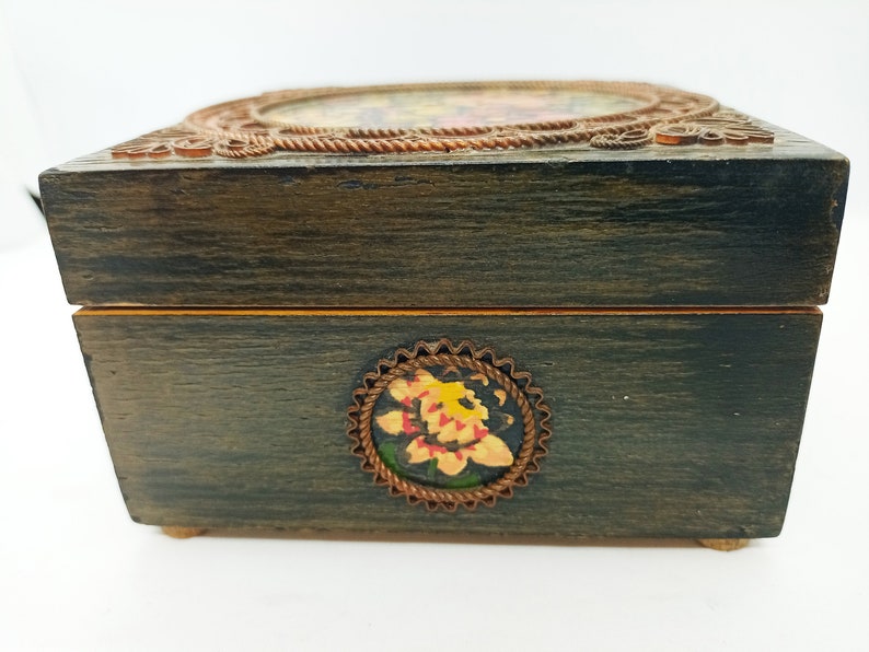 Puede incluir: Una caja de madera con un acabado marr&oacute;n oscuro y un dise&ntilde;o floral en la parte delantera. La caja tiene una tapa con bisagras y un ribete met&aacute;lico decorativo alrededor de los bordes.