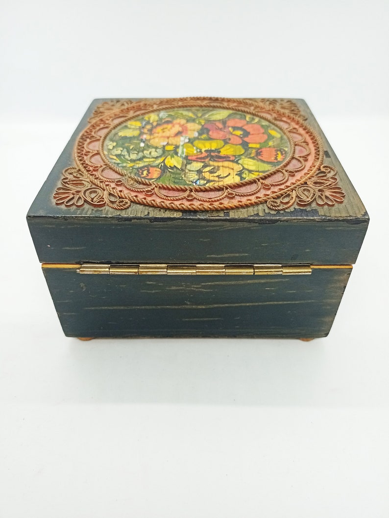 Puede incluir: Una caja de madera verde oscuro con una tapa con bisagras. La tapa presenta un dise&ntilde;o circular con un patr&oacute;n floral en tonos rojos, naranjas, amarillos y verdes. La caja tiene un borde dorado.