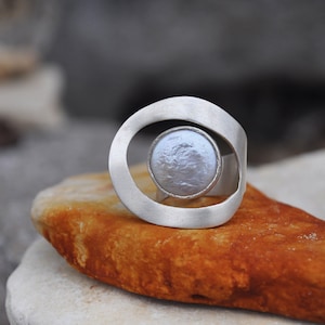 Könnte beinhalten: Ein silberner Ring mit einem großen, runden, weißen Perlmutt, das in einer kreisförmigen Öffnung sitzt. Der Ring liegt auf einer braunen, strukturierten Oberfläche.