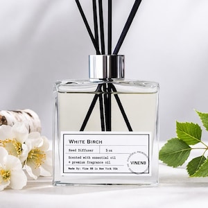 May include: A square glass reed diffuser with black reeds, labeled "White Birch." Birch logs, white flowers, and green leaves are arranged around the diffuser. The diffuser is scented with essential oil and premium fragrance oil.