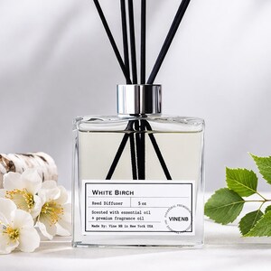 May include: A square glass reed diffuser with black reeds, labeled "White Birch." The diffuser is next to birch logs, white flowers, and green leaves. The product is scented with essential oil and premium fragrance oil. The diffuser contains 5 oz.