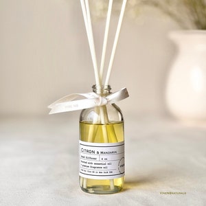 May include: A clear glass reed diffuser bottle filled with yellow liquid and white reeds. The label reads "Citron & Mandarin" and "4 oz". A white ribbon is tied around the neck of the bottle. The text "VINE NB" is on the ribbon.