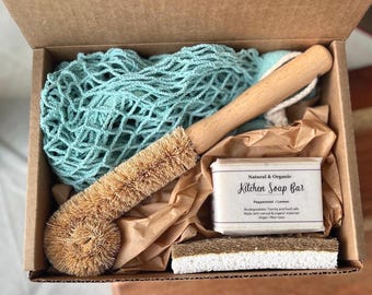 May include: A natural and organic kitchen cleaning set in a cardboard box. The set includes a wooden-handled brush, a green net bag, a soap bar, and a sponge. The soap bar is labeled "Kitchen Soap Bar" with "Peppermint / Lemon" below.