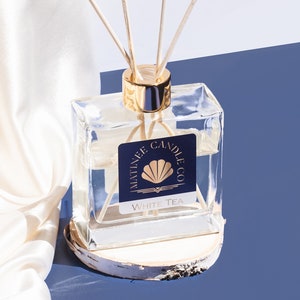 Può includere: Una bottiglia di vetro trasparente con un tappo dorato e bastoncini di legno, riempita di un liquido trasparente. La bottiglia ha un'etichetta bianca con il testo "MATINEE CANDLE CO." e "WHITE TEA". La bottiglia è appoggiata su una piccola base rotonda di legno.