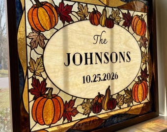 May include: A rectangular stained glass panel in a dark wood frame featuring an oval center with the text The JOHNSONS 10.25.2026. The border is decorated with orange pumpkins, yellow gourds, and multicolored autumn leaves in shades of red and gold.
