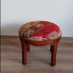 Puede incluir: Un reposapiés redondo de madera con cuatro patas. La parte superior del reposapiés está cubierta con una tela colorida con patrones en tonos rojos, amarillos y marrones.