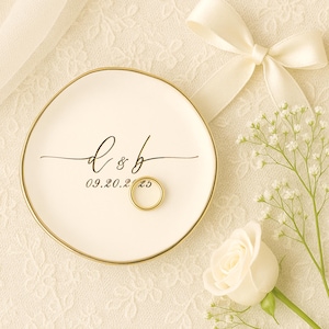 May include: A white ceramic ring dish with a gold rim, featuring the initials "d & b" and the date "09.20.25" in black script. A gold wedding band rests on the dish. The background includes a white rose, baby's breath, and a cream-colored ribbon.
