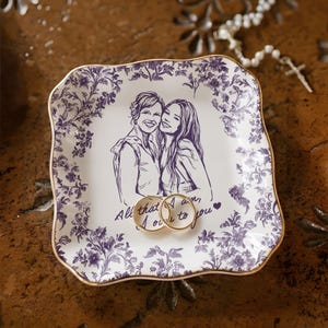 May include: A white ceramic ring dish with a purple floral border and gold trim. The dish features a sketch of two people and the text "All that I am, I owe to you." Two gold rings rest on the dish.