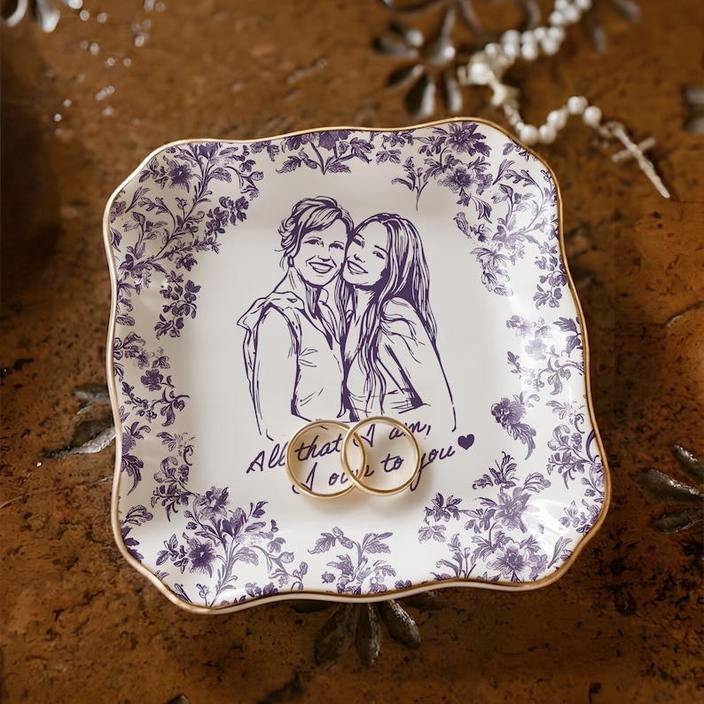 May include: A white ceramic ring dish with a purple floral border and gold trim. The dish features a sketch of two people and the text "All that I am, I owe to you." Two gold rings rest on the dish.
