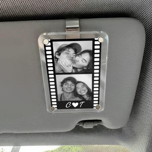 May include: Clear acrylic car visor photo holder with two black and white photos. The photos are in a film strip design. The bottom of the holder has the letters "C" and "T" with a heart symbol between them. The holder is attached to a car visor.