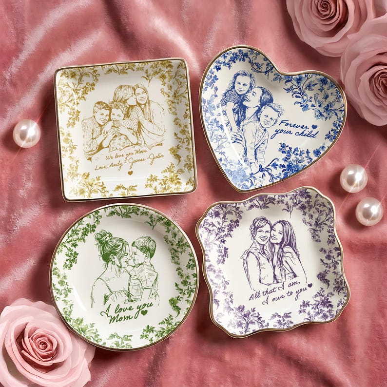May include: Four decorative ceramic plates with gold rims, each featuring a family portrait sketch. The plates are square, heart-shaped, and round, with floral patterns and text: "We love you Mom," "Forever your child," "I love you Mom," and "All that I am, I owe to you."