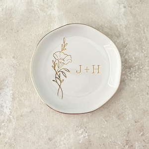 May include: A white ceramic ring dish with a gold floral design and the initials "J + H". The dish has a gold rim and a slightly irregular shape, perfect for holding small jewelry items.