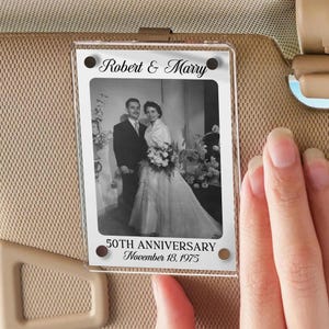 May include: A clear acrylic photo holder with a black and white wedding photo. The text "Robert & Marry" is at the top, and "50TH ANNIVERSARY November 18, 1975" is at the bottom. The holder is attached to a car visor.