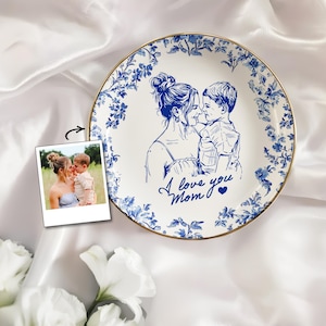 May include: White ceramic plate with a blue floral border and a gold rim. The plate features a blue sketch of a mother and child with the text "I love you Mom". A polaroid photo of the mother and child is next to the plate.