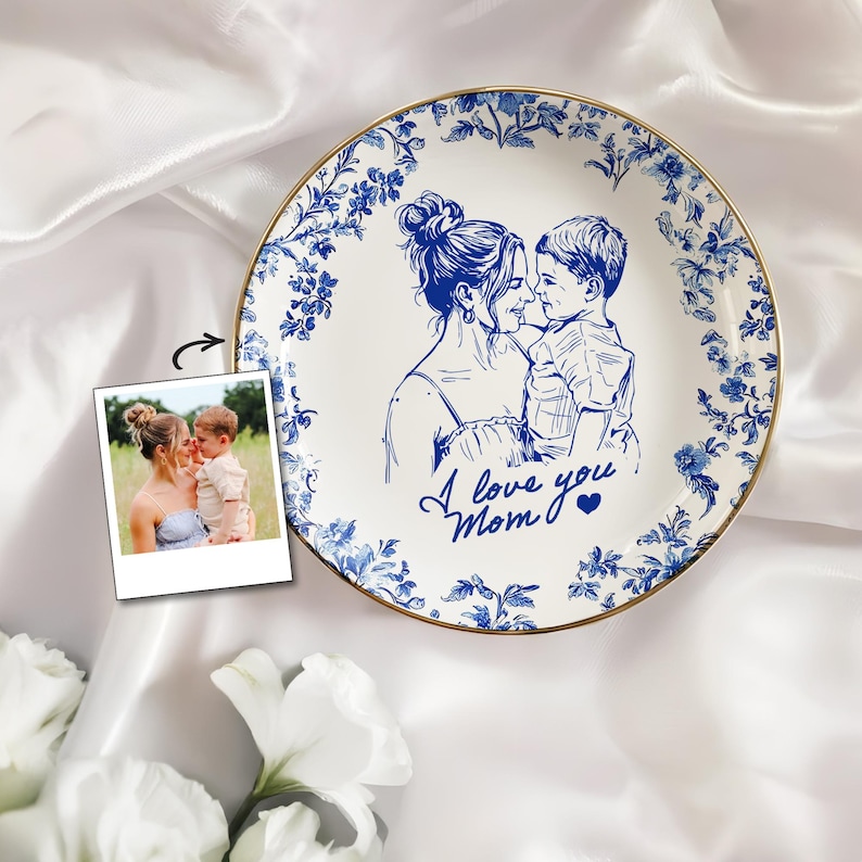 May include: White ceramic plate with a blue floral border and a gold rim. The plate features a blue sketch of a mother and child with the text "I love you Mom". A polaroid photo of the mother and child is next to the plate.