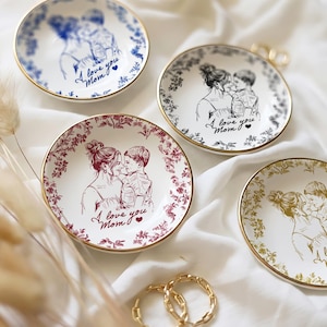 May include: Four small, round decorative plates with gold rims. Each plate features a sketch of a mother and child, with the text "I love you Mom." The plates have different color schemes: blue, black, red, and gold, with floral or line art borders.