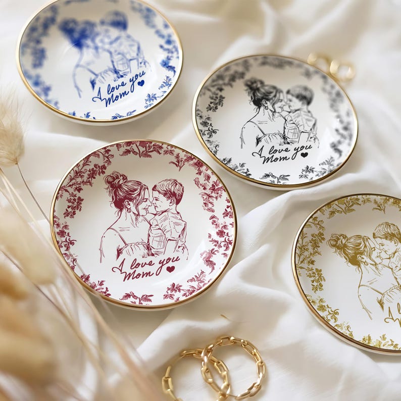 May include: Four small, round decorative plates with gold rims. Each plate features a sketch of a mother and child, with the text "I love you Mom." The plates have different color schemes: blue, black, red, and gold, with floral or line art borders.