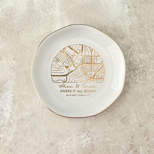 May include: A white ceramic ring dish with a gold map design. The dish has a gold rim and features the text "Alex & Sara WHERE IT ALL BEGAN 08.20.2020 | Phoenix, AZ". The map design is in gold.