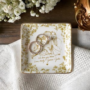 May include: Square, white ceramic trinket tray with a gold floral design and a gold rim. The tray features a family portrait and the inscription "We love you Mom, Love, Andy - James - Julia." Two rings rest on the tray.