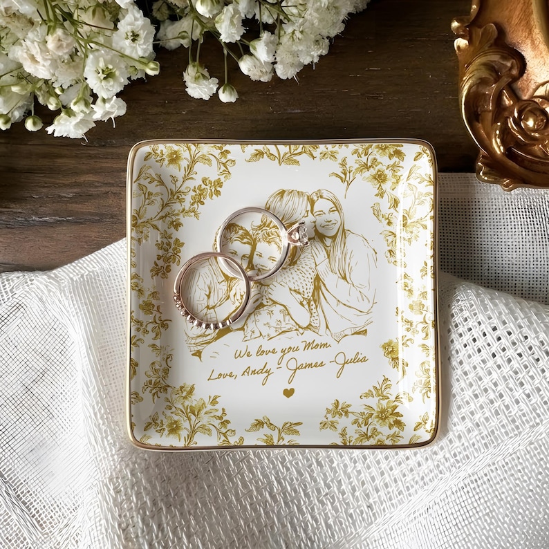 May include: Square, white ceramic trinket tray with a gold floral design and a gold rim. The tray features a family portrait and the inscription "We love you Mom, Love, Andy - James - Julia." Two rings rest on the tray.