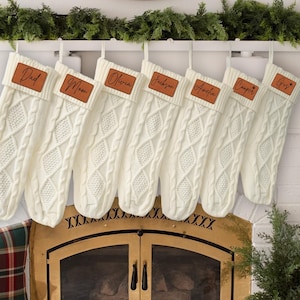 May include: A collection of seven cream-colored Christmas stockings hanging from a white mantel. Each stocking has a cable knit design and a brown leather patch with a name on it. The names include "Dad", "Mom", "Olivia", "Jackson", "Amelia", "Cooper", and "Gus".