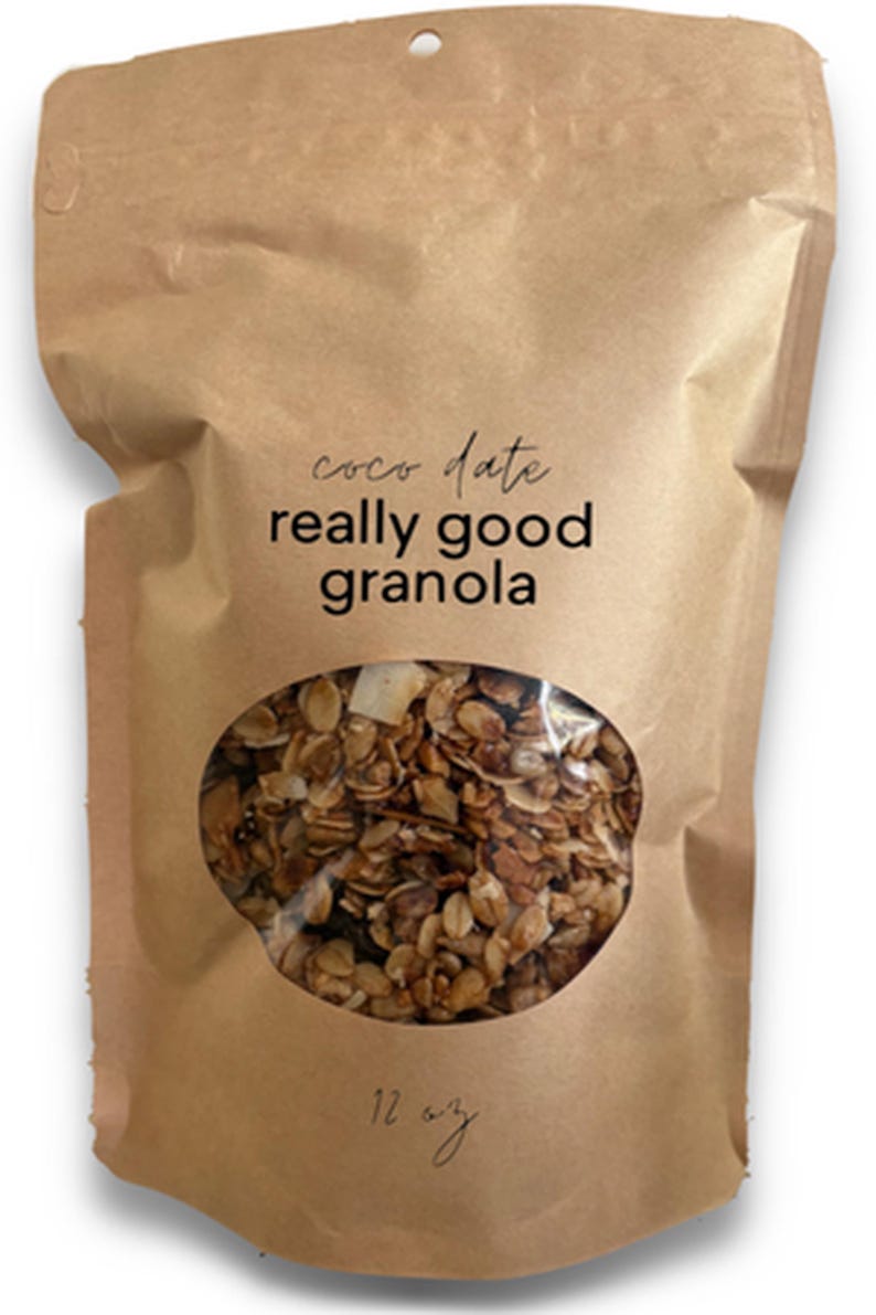 K&ouml;nnte beinhalten: Ein brauner Standbeutel aus Papier mit Granola, der ein ovales Sichtfenster besitzt, durch das der Inhalt zu sehen ist. Der Aufdruck auf der Vorderseite lautet "coco date really good granola" und "12 oz" in schwarzer Schrift.