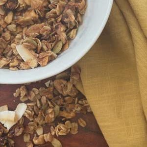 K&ouml;nnte beinhalten: Eine wei&szlig;e Sch&uuml;ssel gef&uuml;llt mit Granola aus ger&ouml;steten Haferflocken, Kokosflocken, K&uuml;rbiskernen und Trockenfr&uuml;chten. Loses Granola ist auf der dunklen Holzoberfl&auml;che neben der Sch&uuml;ssel verstreut, begleitet von einem gefalteten senfgelben Leinentuch.